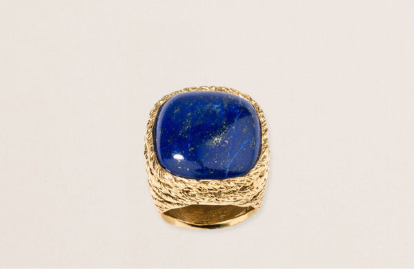 The Lapis Miki Ring by Aurelie Bidermann showcases a vibrant large polished lapis stone with a subtle marbled look set on a textured gold-plated band. The striking color of the stone contrasts beautifully against its plain light gray backdrop for a unique style.