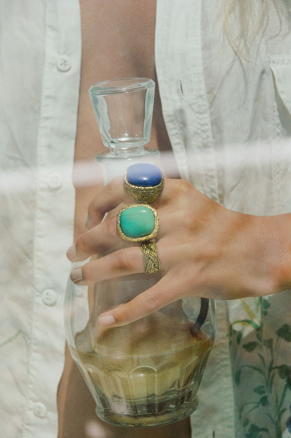 A person in a white shirt holds a stopper glass bottle filled with light brown liquid. On their hand are two rings—one is the Aurelie Bidermann Lapis Miki Ring featuring blue and turquoise stones, and the other is a striking yellow gold-plated ring.